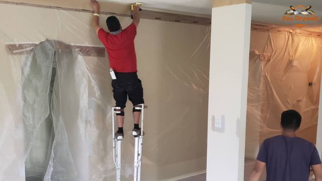 Contractor on stilts installing or taping drywall near the ceiling while another worker prepares the room, with walls and openings covered in protective plastic for renovation.
